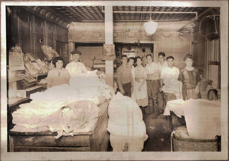 An old black and white photo with a young couple standing on the left behind a table piled with white fabric for the laundry and a group of seven people, some young and some old who are the employees standing in the back of the room. They are all faintly smiling.