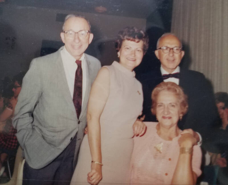 An old color snap shot of two older couples dressed up and at a fancy banquet. The couple on the left are standing, the man is wearing a gray suit jacket with a white shirt and red tie,  and the woman is wearing a light pink sleeveless dress with a high collar. The couple to the right, the woman is sitting at a table and has on a pink dress with a pink short sleeved cardigan, and the man standing behind her has on a black tuxedo.