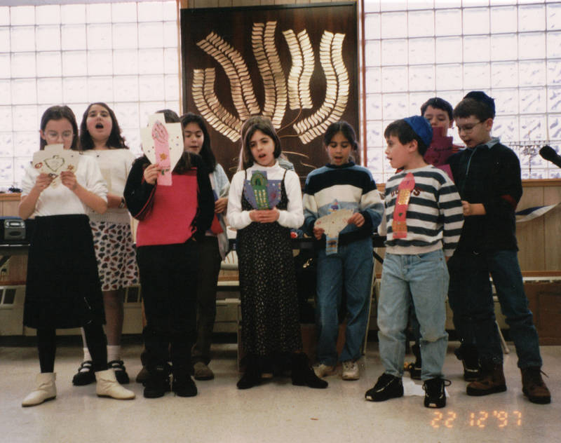 A group of children  8 to 9 years old are singing in front of lit up windows and there is  a plaque behind them with brass plates shaped like leaves rising up in wavy branches.