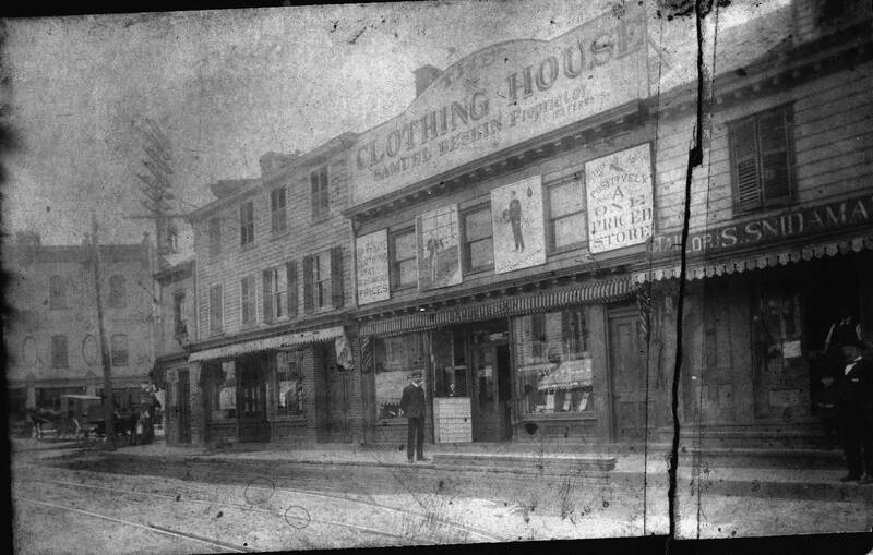  A very old black and white photo that was cut along the right side and has been pieced together. The image depicts a street with trolley tracks, and a row of wooden buildings. The middle building has a large sign at the top of its edifice that says “The Clothing House, Samuel Beskin, proprietor.” A young man, who is Samuel Beskin, stands in front of his store’s front door in a dark suit with a newsboy cap and a mustache. The building to the right of this has a smaller sign directly over the door and extending the length of the building that says “Tailor: S. Snidaman.” The owner, Samuel Snidaman, stands in the lower right hand corner in front of his front door, wearing a black tuxedo, black silk top hat and a mustache.