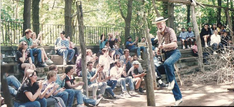 A tall man with a straw hat and a white beard is singing and skipping while playing a banjo. Young people sit on concrete bleachers and are clapping and singing along.