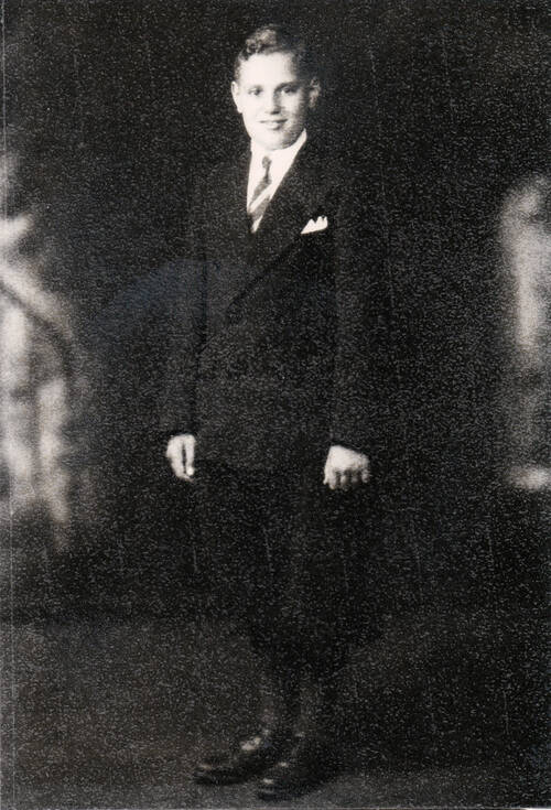 An old grainy black and white photograph of a boy around 13 years old, wearing a black suit jacket with a white collared shirt, a striped tie, and a white handkerchief folded  in his breast pocket. He is also wearing dark knickerbocker shorts, dark socks and shiny dark leather shoes. The portrait was taken in a photography studio with blurry stained glass window shapes in the background. He is smiling slightly into the camera and his short hair is parted to the side