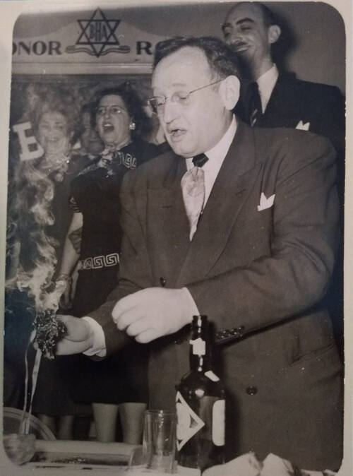 A man stands at a table looking down and singing with a burning bunch of incense in his hand and a bottle of wine in front of him. There are women behind him singing and a banner above them that has the initials B.H.A. on it for Beacon Hebrew Alliance. It is black and white and the clothing of the people suggests it is the 1950s.