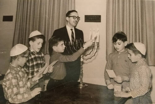 A black and white photo of a man wearing a black suit and tie, with a mustache and dark framed glasses, and a dark kippah on his head is singing a blessing behind a Hanukkah menorah on a table. The candles in the menorah are being lit by a small boy wearing a kippah. Four other boys flank the man and the boy lighting the candles. They are all wearing kippahs and singing while reading the blessing from papers in their hands.