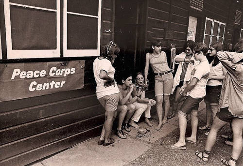 A group of teenage girls are standing and sitting in the doorway of a dark wooden building. There is a dark banner with the words “Peace Corps” in white letters hanging under the window on the outside of the building.