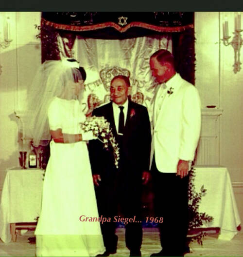 A short older man in a suit and tie stands between a bride in white and a groom in a white suit jacket. They are all on a bimah with an ark behind them that has a silver, shiny curtain with a crown embroidered on it. A caption on the photo says: Grandpa Siegel … 1968.