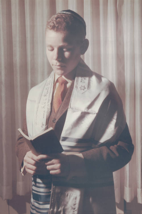 A thirteen year old boy wearing a yarmulke and a white tallis looks down at a bible in his hands. He is standing in front of a velvet curtain.