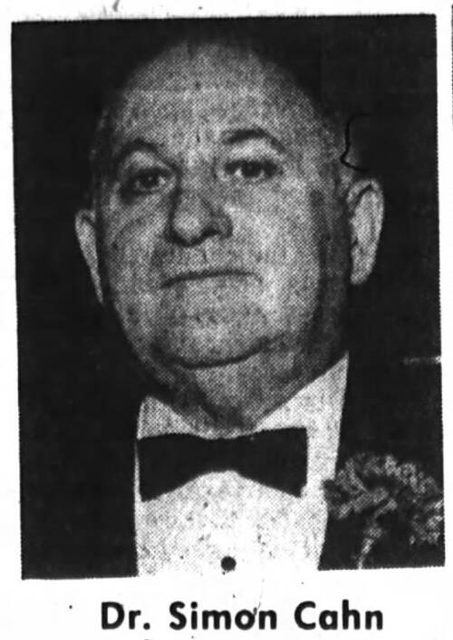 A black and white photo of an elderly man standing, wearing a black tuxedo with a bow tie and a flower corsage. He has a bald head and heavy jowls. He is smiling slightly at the camera.