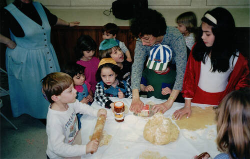 A color photo of children rolling dough on a table with a large ball of dough in the center. A woman stands behind one of the small children, helping them to roll it out. Many children stand near her, waiting their turn at the activity. An older girl in a red velvet jacket and skirt stands to the right of the woman, and is looking on, her dough is rolled out in front of her.
