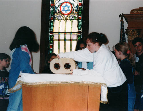 Two teenage girls stand to either side of a wooden lectern, where a large white Torah school is laid on it. They are on a bimah and light shines through a tall 