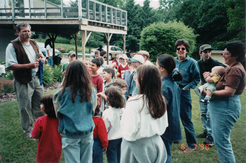 A color photo outside with a group of children and adults looking at a man in a brown leather vest and white t-shirt. He has just performed a magic trick for them.