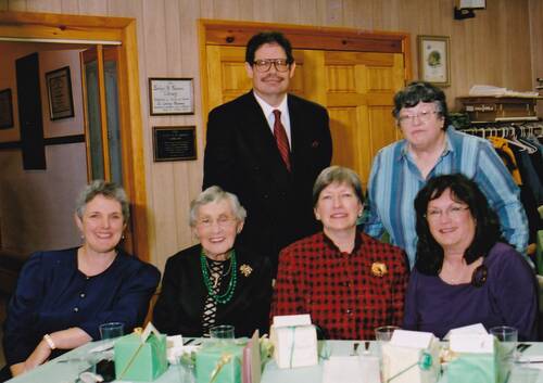 A color photo of a group of people, four women sitting, and one man and a woman standing behind them. They are at a table covered in sea foam green and white party boxes. The women sitting in front are all wearing dresses and range in age from 40s to 90s. The man in the back is wearing a black suit and red tie, the woman next to him is wearing a blue striped button down shirt. They are all smiling.