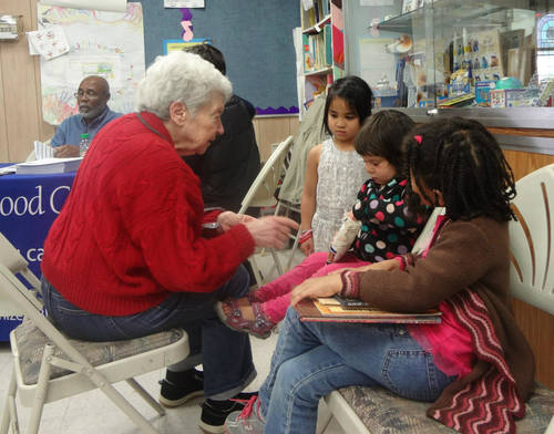 An old woman with white hair and a bright red sweater is seated across from a group of small girls of various races. A table is behind them with a banner for a Blood drive and a man sitting at it.