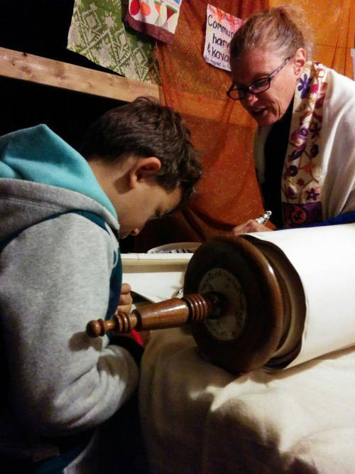 : A young boy is seated looking at a large Torah Scroll open in front of him. A woman in a prayer shawl stands across from him and is speaking to him.