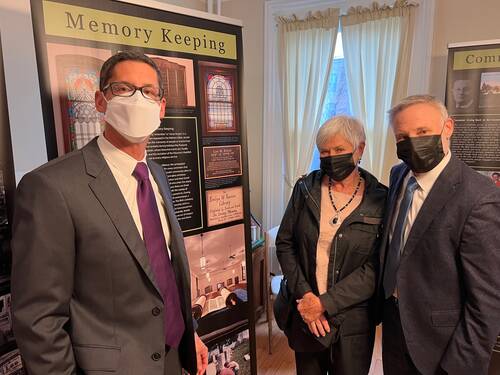 Three adults wearing face masks look at the camera. The man on the left is tall with dark hair, dark glasses, a white face mask, and a dark gray suit jacket with purple tie. A woman with white short hair, a black face mask, white blouse and black jacket stands next to another man with white short hair, black face mask, dark grey suit jacket and blue tie. A black panel is behind them with colorful photos.