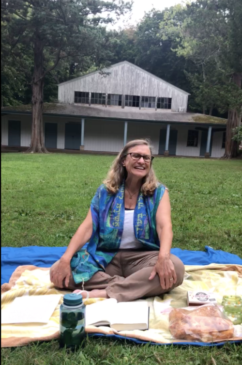  A woman with blond hair, wearing glasses, and a blue prayer shawl  smiles and sits cross-legged on a blanket in the grass. She has a book open in front of her and a loaf of braided challah bread. There is a large white wooden barn-type building behind her.