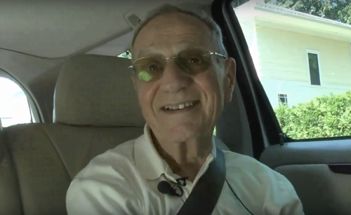 An elderly man is sitting in a car with a seat belt on. He is smiling and looking at the camera. He is wearing a white collared polo shirt, a lavalier microphone clipped to his collar, and tinted glasses.