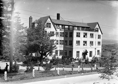 A black and white photo of a large Arts and Crafts style hotel building with the letters HOTEL NITGEDAIGET on its face. Old style Model T cars are parked in front. Rolling hills are in the background.