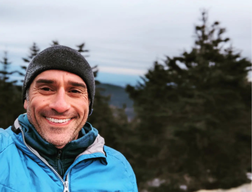 A selfie photo taken on top of a mountain with dark green conifer trees and bald rocks. The man is standing and smiling at the camera. He is wearing a black beanie that is frosted, and a blue shell jacket with a darker blue fleece lining. His skin is olive colored, and he has light gray stubble where a beard would be.