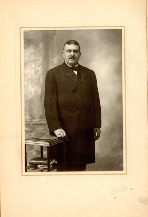  A sepia toned black and white portrait of a large man, standing next to a small table with books under it. He is wearing a long dark, double breasted suit jacket over dark trousers, and a white collared shirt with a black bow tie. His brown hair is parted down the middle and he has a thick dark mustache. He is looking over the camera into the distance with a serious expression.