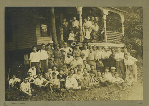 An old photograph in sepia tone of a group of young men and boys sitting and standing in front of a white house with a high lattice porch. There are trees all around the scene, and some of the boys are hanging off the porch. They all are wearing white shirts and many have hats and walking sticks.